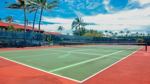 Een buiten tennisbaan met een rode ondergrond, omgeven door een hek, palmbomen en een gebouw met een rood dak.