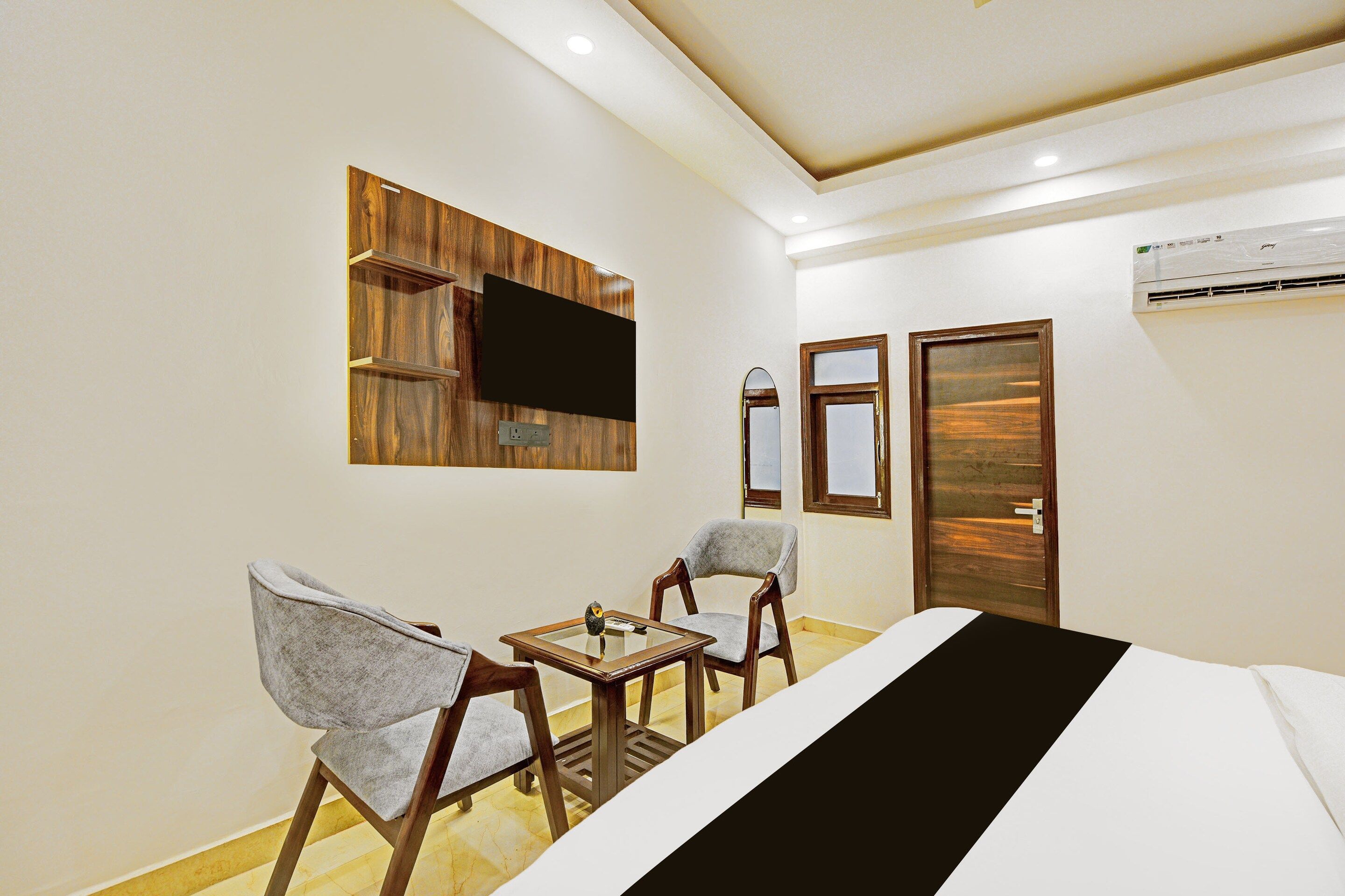 A hotel room with a bed, two chairs, a small table, a flat-screen TV mounted on a wooden panel, a door, and an air conditioning unit.