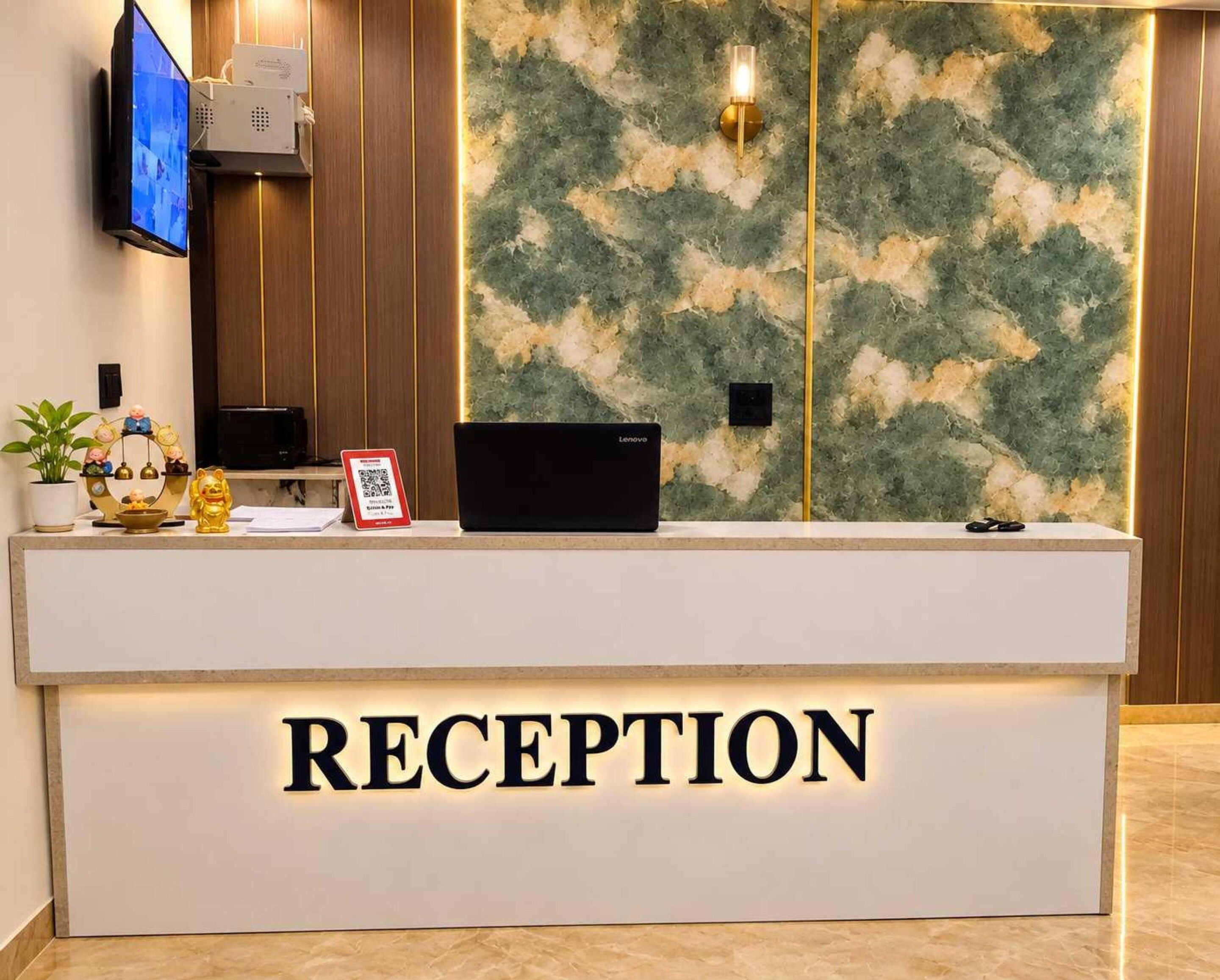A reception desk with a 'RECEPTION' sign, a flat-screen TV, a Lenovo laptop, a small plant, and a decorative item.