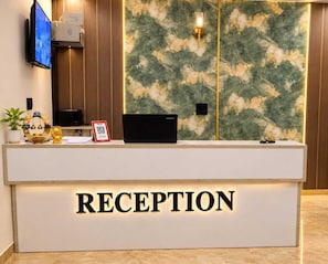 A reception desk with a 'RECEPTION' sign, a flat-screen TV, a Lenovo laptop, a small plant, and a decorative item. - Classic Charmwood Inn (Faridabad)