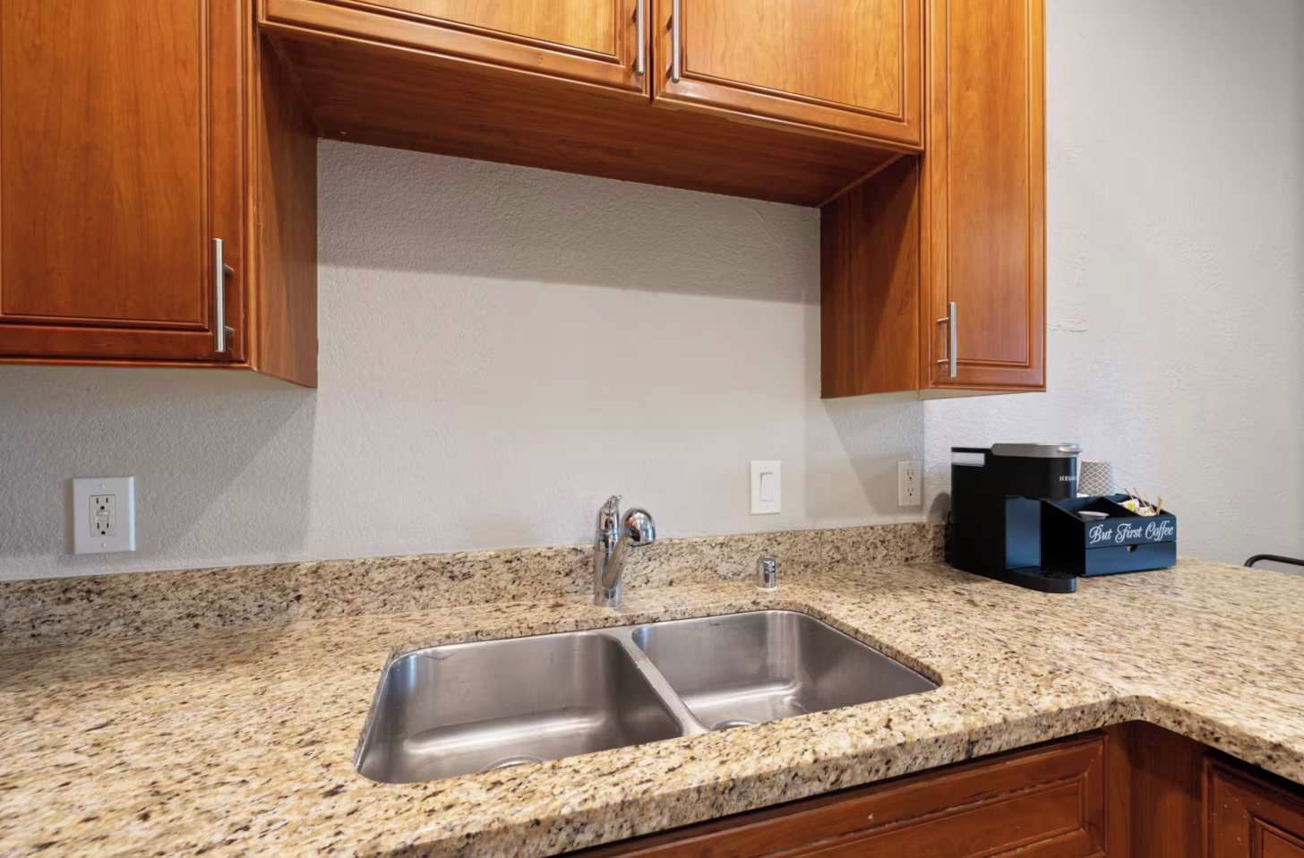Una cocina con encimeras de granito, pileta de acero inoxidable, muebles de madera y una cafetera sobre la encimera.