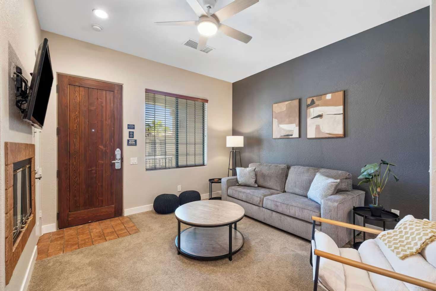 A living room with a grey sofa, a round coffee table, a TV mounted on the wall, a ceiling fan, and a window with blinds.