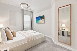 Un dormitorio con cama, una mesita de noche con lámpara, un espejo grande, un cuadro de playa y una ventana con persianas.