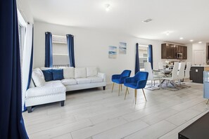 Ein modernes Wohnzimmer mit einem weißen Sofa, blauen Sesseln, einem Essbereich mit Tisch und Stühlen sowie einer Küche im Hintergrund.