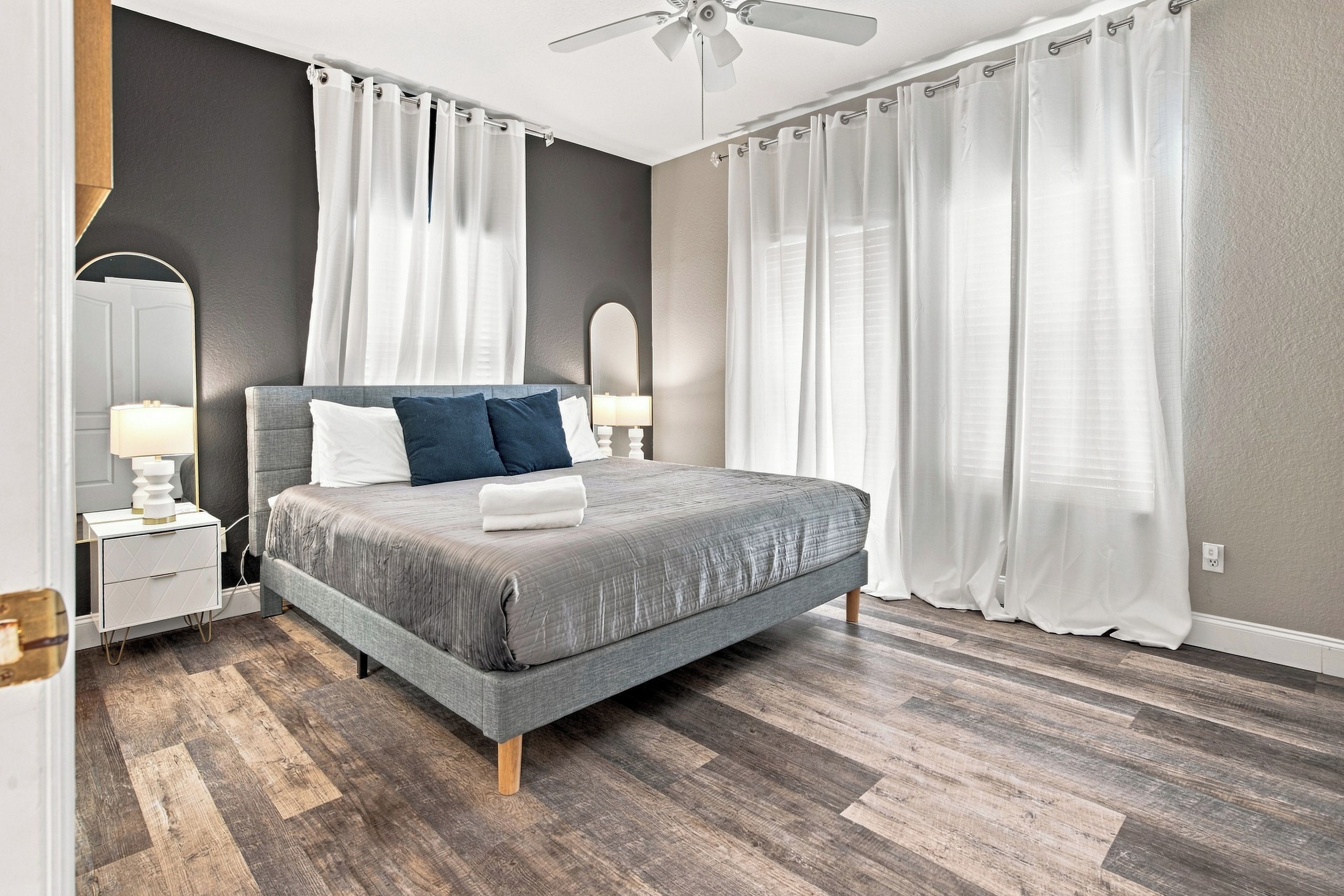 Ein modernes Schlafzimmer mit einem grauen Bett, weißen Vorhängen, einem Deckenventilator und einem Nachttisch mit einer Lampe.