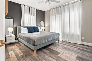 Un dormitorio moderno con cama gris, cortinas blancas, ventilador de techo y mesita de noche con lámpara.