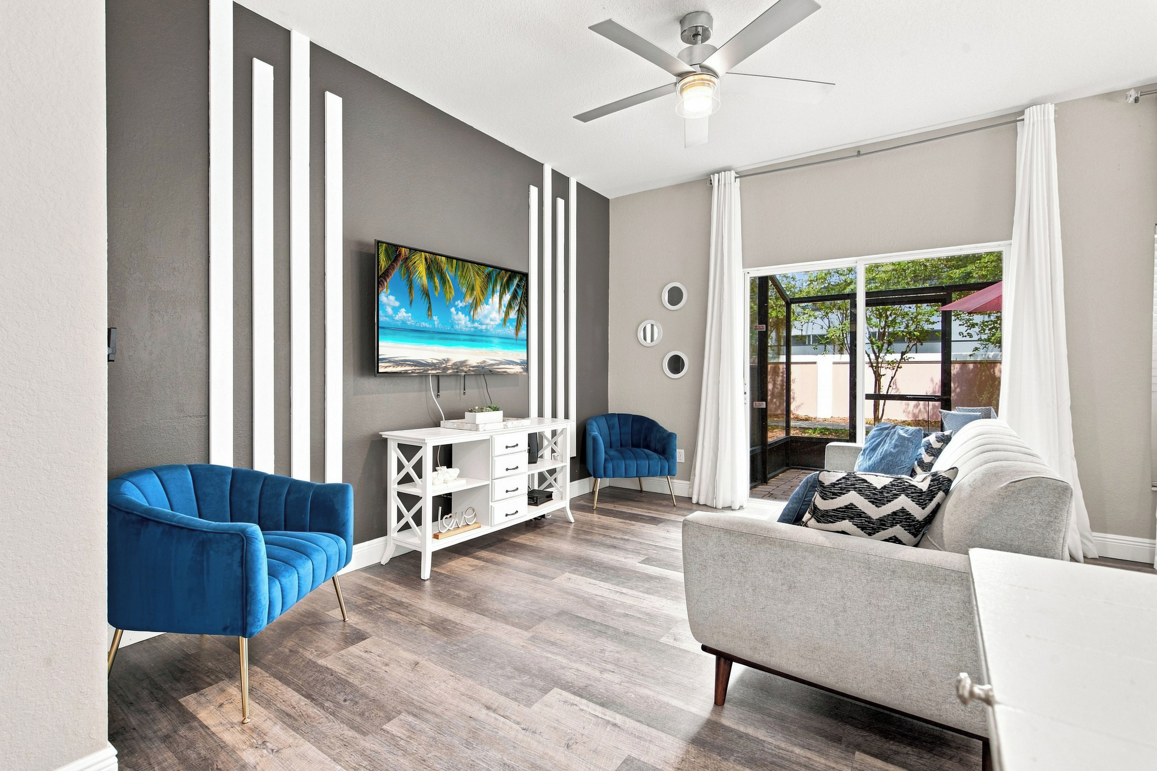 A modern living room with a flat-screen TV, two blue armchairs, a grey sofa, and a ceiling fan.
