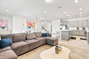 Un saló modern amb un sofà seccional, una taula de centre i una zona de cuina amb armaris i electrodomèstics.