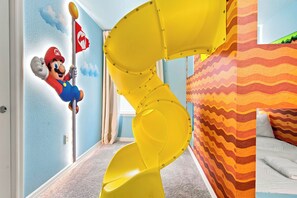 A room with a yellow spiral slide, a Mario character decoration, and a bed with a patterned bedspread. - Kids Slide Room, Arcade & Game Room, Sleeps 24 (Clermont)