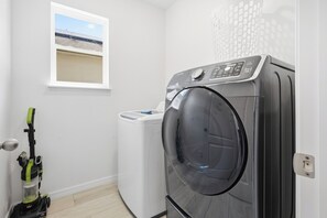 Een kleine wasruimte met een voorlader wasmachine en droger, een raam met uitzicht op een gebouw, en een stofzuiger.