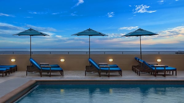 Zona de piscina amb gandules i ombrel·les, una vista despejada de l'oceà i un solàrium amb rajoles.