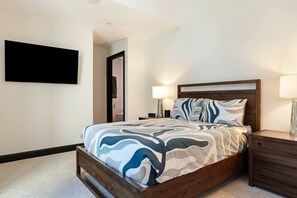 Ein Schlafzimmer mit einem Bett, Nachttischen, einem an der Wand montierten Fernseher und einer Tür zu einem anderen Raum.