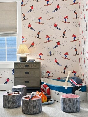 Ein Kinderzimmer mit ski-bezogenen Tapeten, einer Lampe, einer Kommode und einem Bett mit blauer und weißer Bettwäsche.