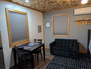 A room with a dining table, chairs, a black sofa, a clock, and a window with blinds. - Cabin Harper, 1-bedroom cabin in the heart of Amish Country in charming Clare (Clare)