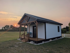 A small, single-story building with a porch, a door, and a window, situated in a grassy field with a sunset in the background. - Cabin Harper, 1-bedroom cabin in the heart of Amish Country in charming Clare (Clare)