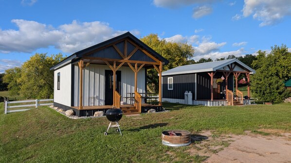 A rustic cabin with a porch and a smaller attached structure, both with wooden beams and metal siding, set in a grassy area with trees in the background.
