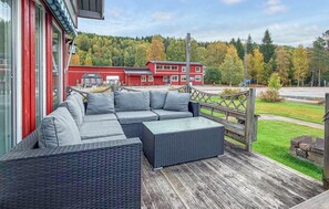 Ein überdachter Außenbereich mit Rattanmöbeln, darunter ein Sofa und ein Tisch, mit Blick auf eine bewaldete Gegend und ein rotes Gebäude.