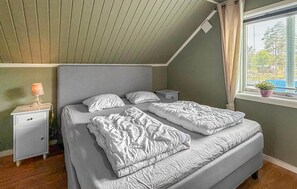 Ein Schlafzimmer mit schrägem Dach, einem Bett mit zwei Kissen, einem Nachttisch mit Lampe und einem Fenster mit Jalousien.