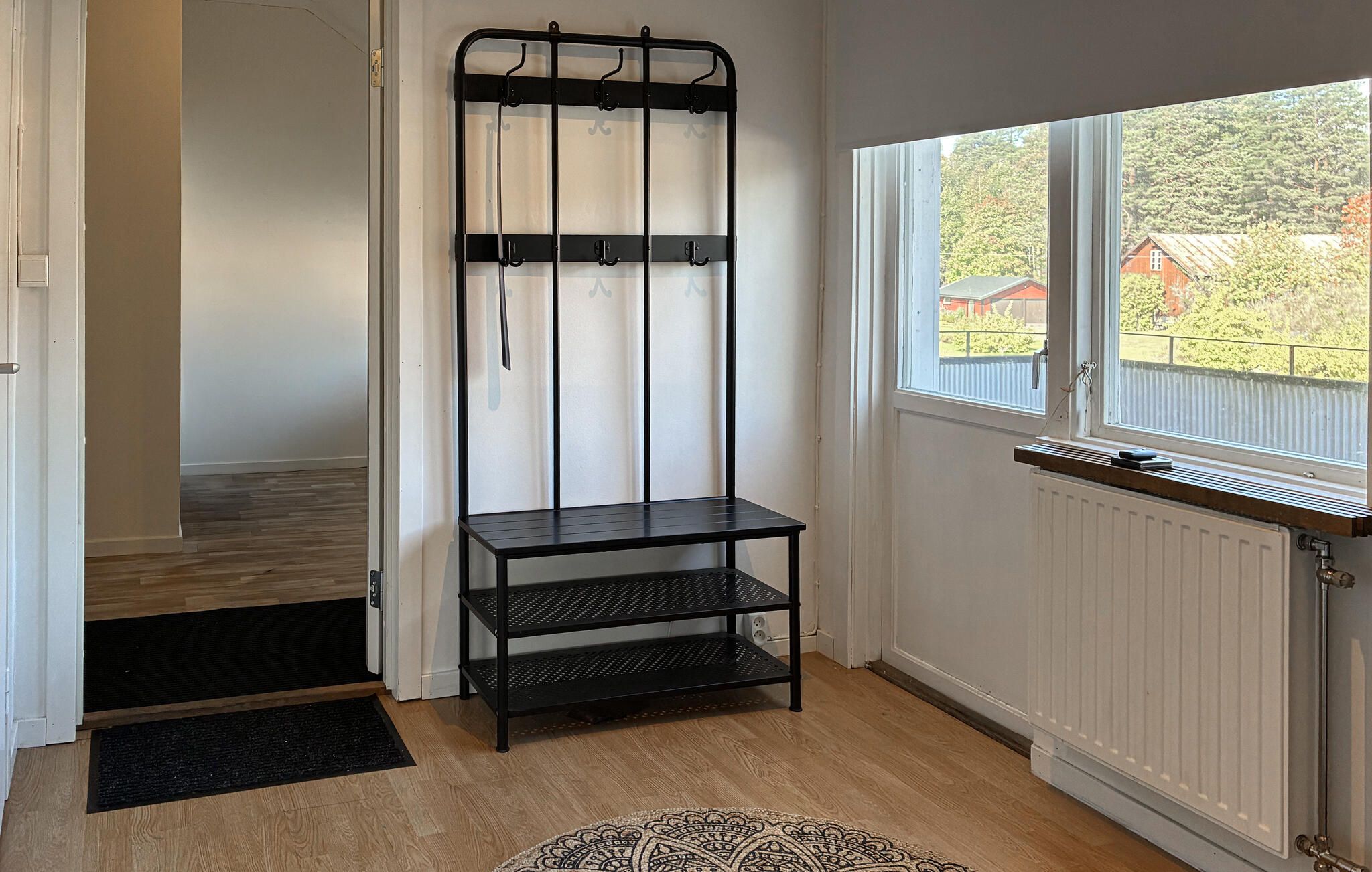 Huoneessa on musta metallinen hyllykkö, ikkuna, josta on näkymä vehreään maisemaan, ja patteri.