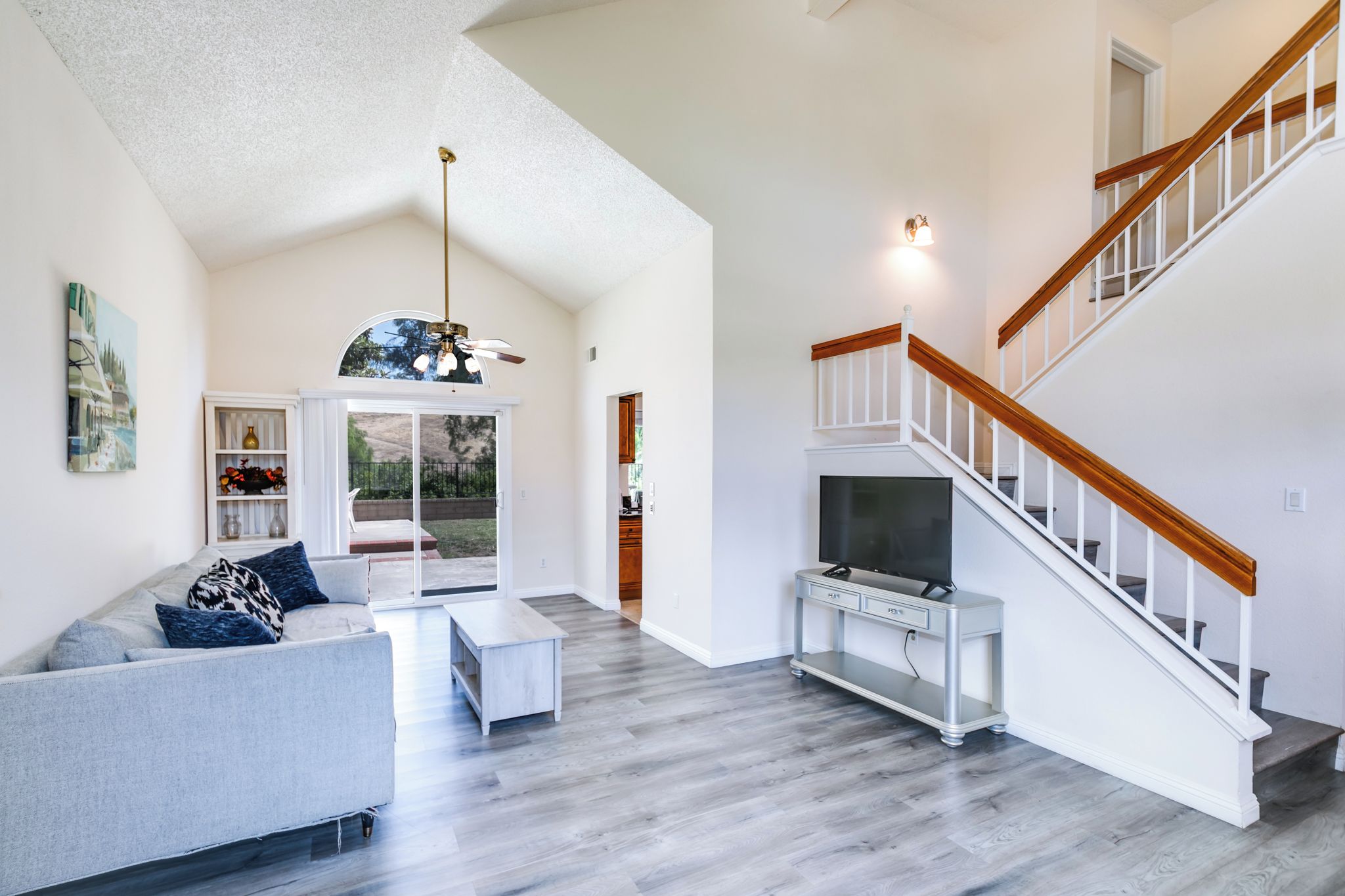 A spacious living room with a grey sofa, a coffee table, a TV on a stand, and a staircase with wooden railings.