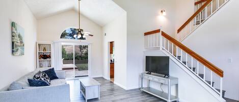 A spacious living room with a grey sofa, a coffee table, a TV on a stand, and a staircase with wooden railings.