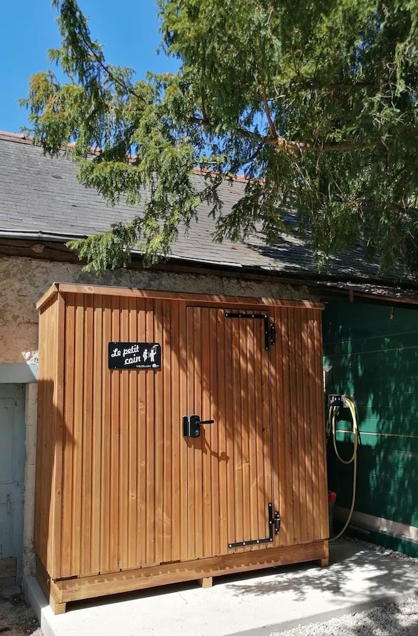 Een houten tuinhuisje met een bord waarop 'Le petit coin' staat en een tuinslang aan de zijkant.