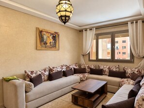 A living room with a sofa set, a coffee table, a painting on the wall, and a window with curtains. - Guéliz - Stylish Apartment with Balcony (Marrakesh)