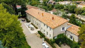 A two-story building with a tiled roof, a paved courtyard with outdoor furniture, and a river in the background. - Exclusive Estate - La Chapelle aux Acacias (Caucalières)