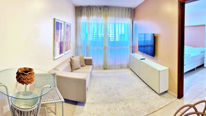 A modern living room with a sofa, a glass coffee table, a TV on a stand, and a bed in the background. - Vacanze - Latitude 10 (Maceió)