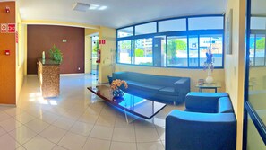 A hotel lobby with a reception desk, a sofa, a coffee table, and a large window offering a view of the outside. - Vacanze - Latitude 10 (Maceió)
