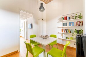 Una habitación luminosa con una mesa blanca, sillas verdes y una estantería con libros y plantas.