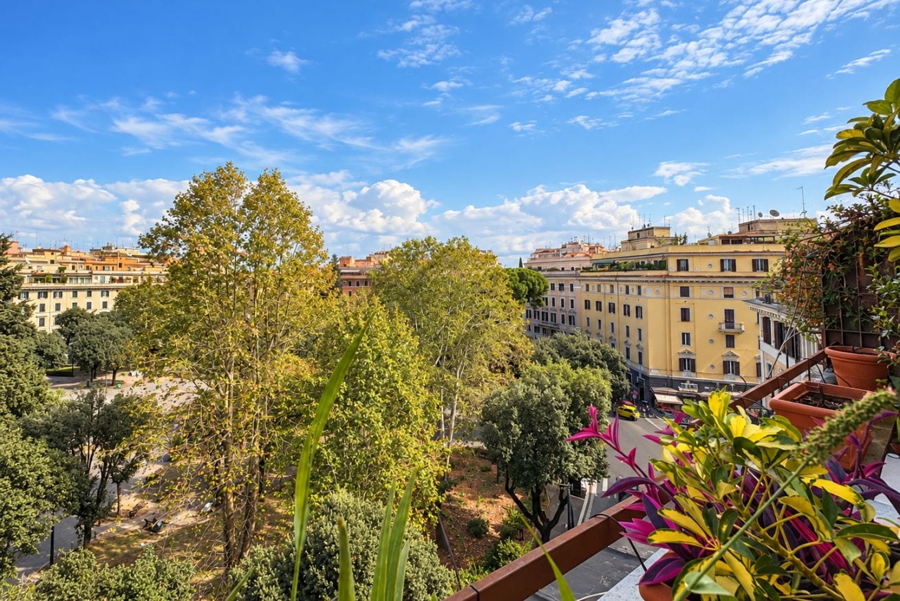 A balcony with potted plants, overlooking a cityscape with buildings and trees.