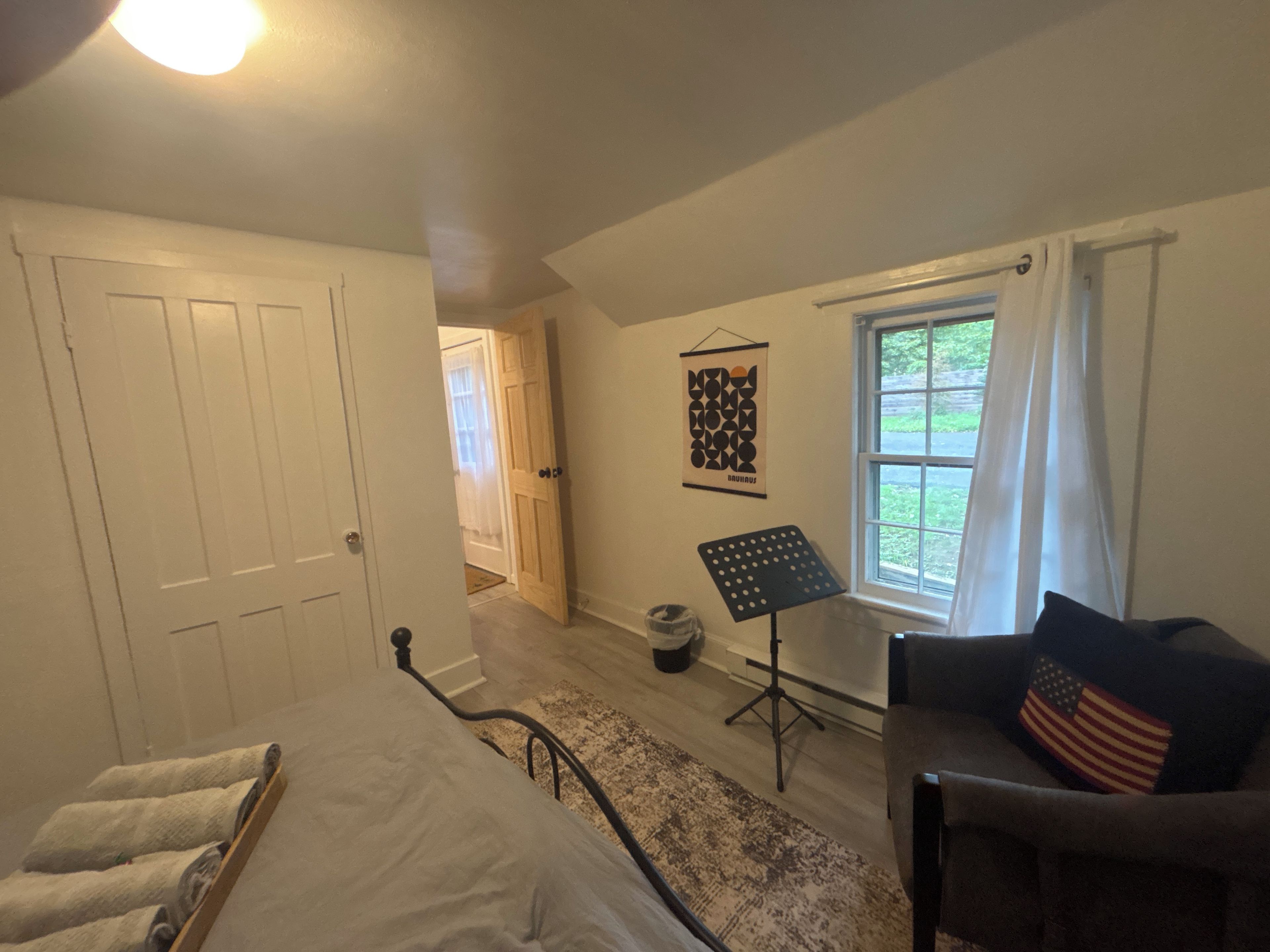 Ein Schlafzimmer mit einem Bett, einem Sessel mit Kopfkissen mit amerikanischer Flagge, einem Notenständer und einem Fenster mit Vorhängen.
