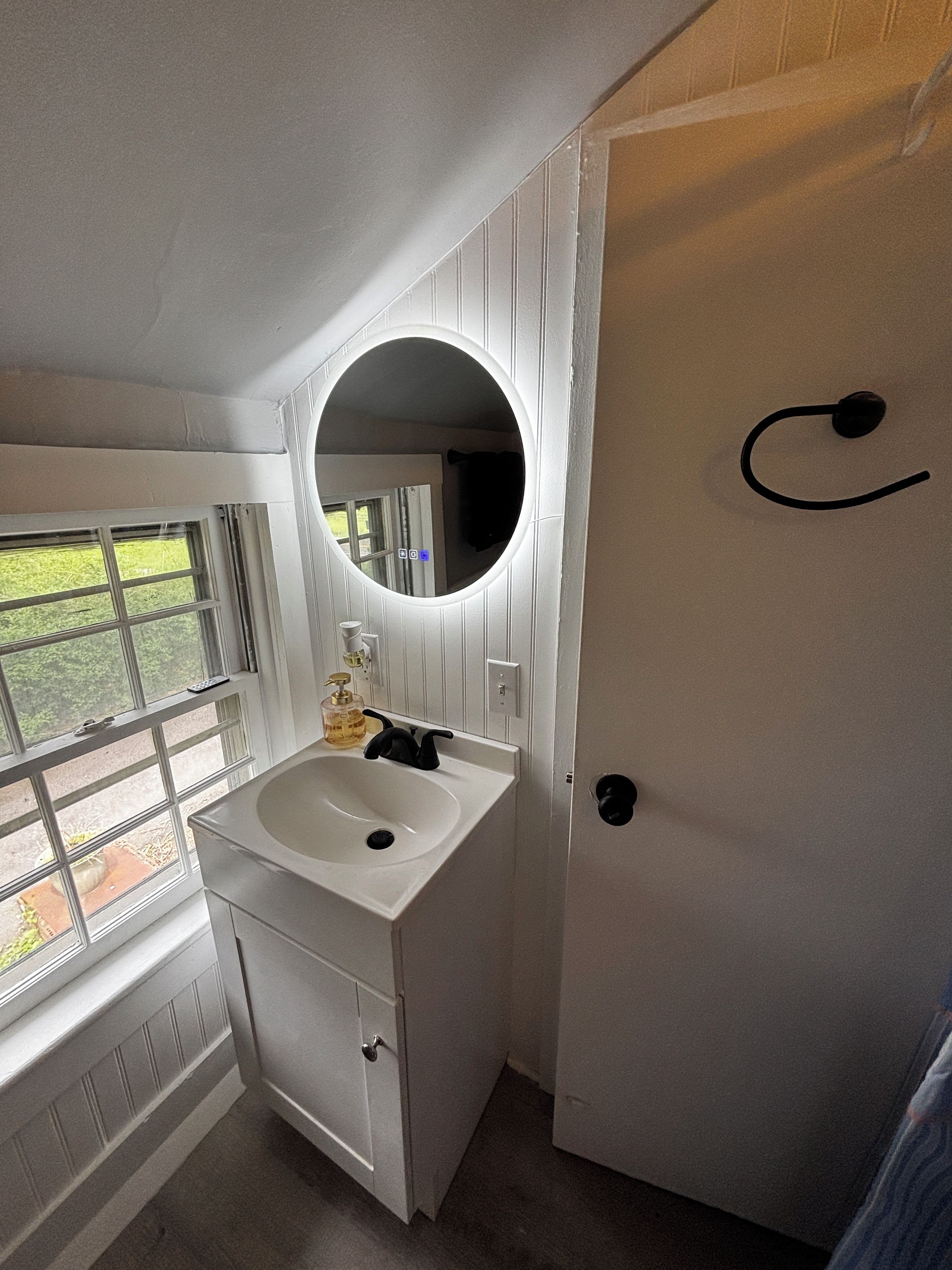 Un baño con tocador blanco, espejo redondo, ventana y puerta con pomo negro.