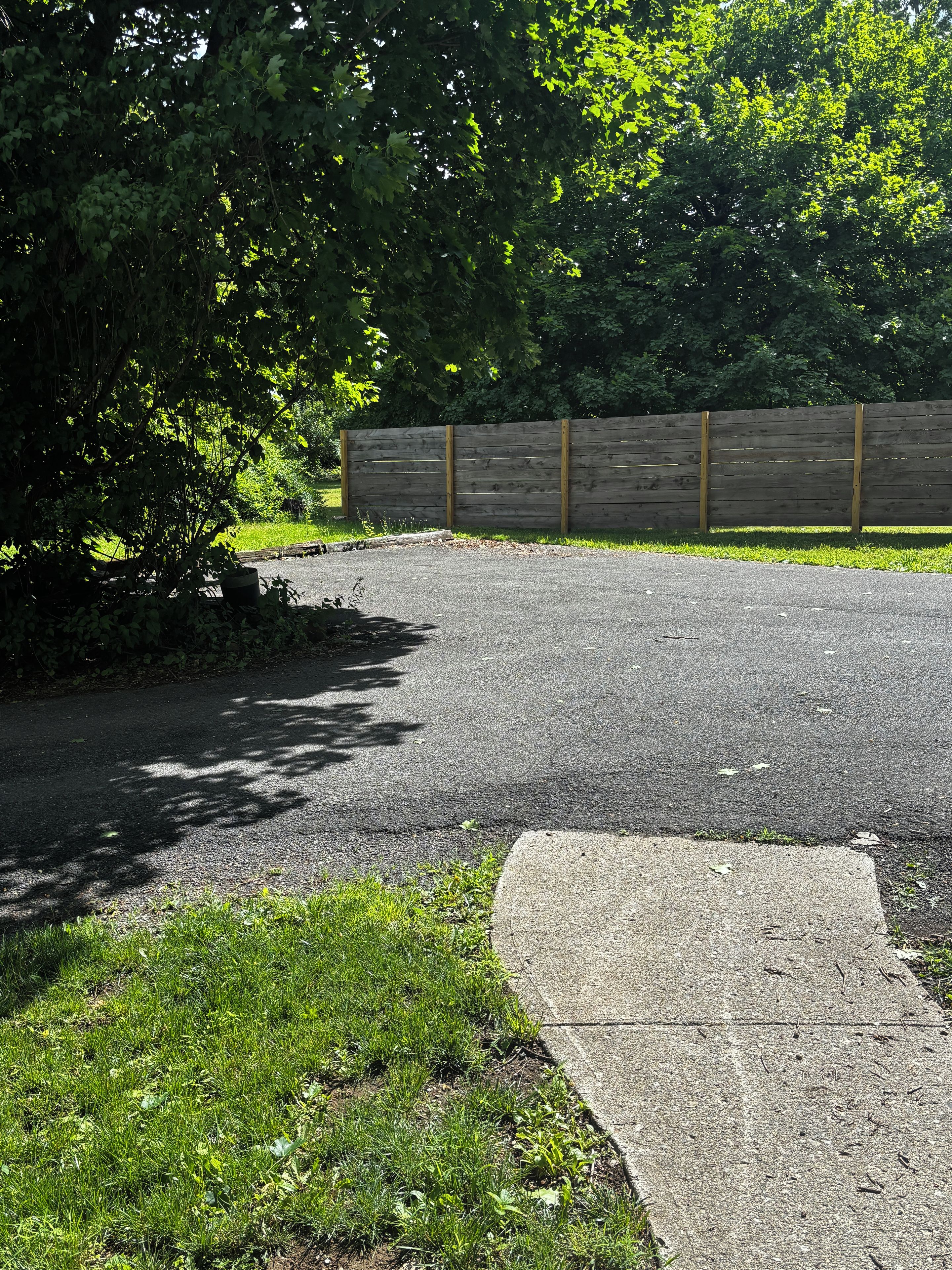 A paved driveway leading to a wooden fence, surrounded by greenery and a concrete path.