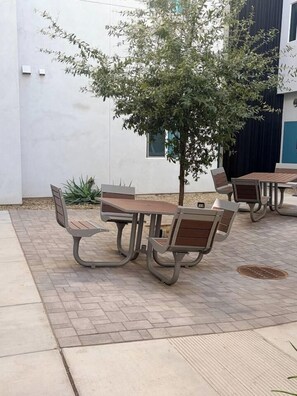 Un espacio al aire libre con mesas y sillas, un árbol y un edificio al fondo.