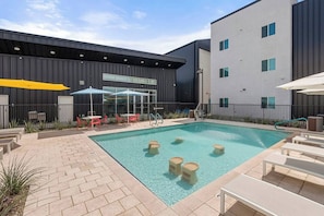 Ein moderner Poolbereich im Freien mit Liegestühlen und Sonnenschirmen, umgeben von einem Gebäude mit dunkler Fassade und großen Fenstern.