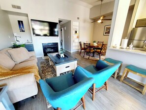 Ein modernes Wohnzimmer mit einer Couch, zwei blauen Sesseln, einem Couchtisch und einem Fernseher. Es gibt einen Essbereich mit Tisch und Stühlen sowie eine Küche mit Kühlschrank und Spüle.