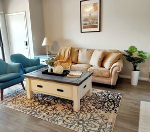 Ein Wohnzimmer mit einem beigen Sofa, einem blauen Sessel, einem Couchtisch, einem gemusterten Teppich und einem gerahmten Bild an der Wand.
