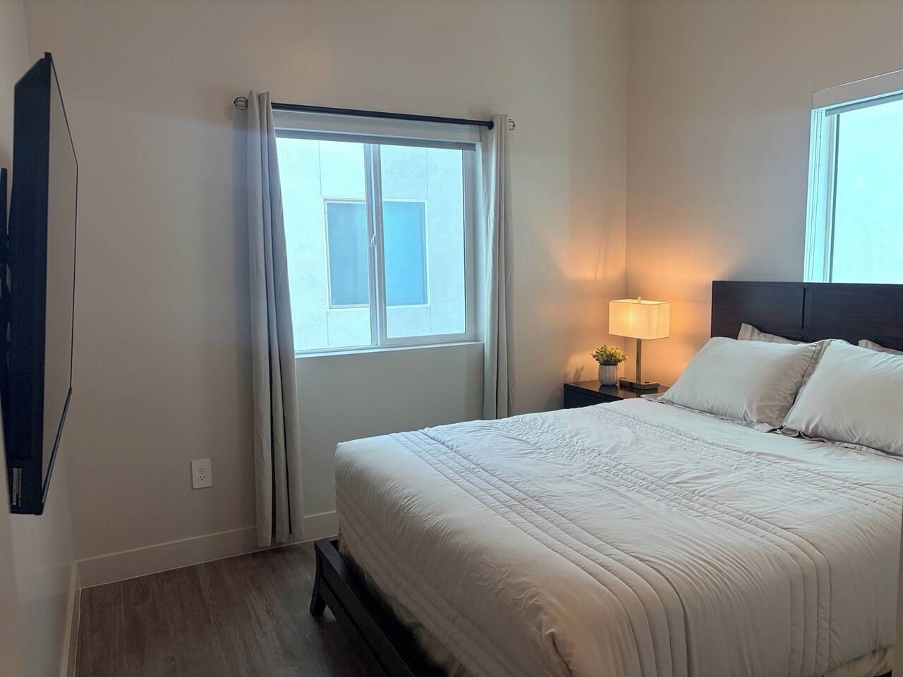 A hotel room with a bed, a nightstand with a lamp, a window with curtains, and a wall-mounted TV.