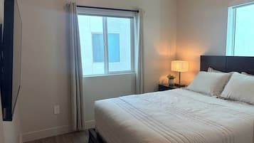 A hotel room with a bed, a nightstand with a lamp, a window with curtains, and a wall-mounted TV.