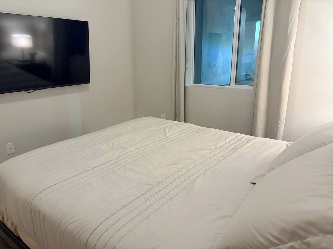 A bedroom with a bed, a flat-screen TV mounted on the wall, and a window with curtains.