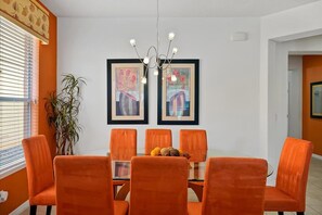 Een eethoek met een glazen tafel, omringd door zes oranje stoelen, kunstwerken aan de muur en een hangende lamp.