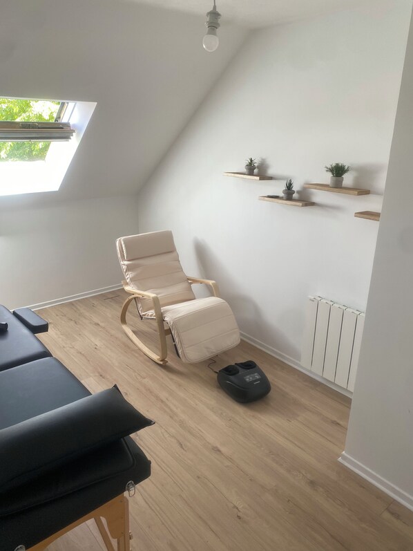 Una habitación con techo inclinado, una silla de masaje, una camilla de masaje, suelo de madera, un radiador y estantes en la pared con plantas en macetas.