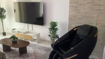 Uma sala de estar moderna com televisão de tela plana, poltrona de massagem, mesa de centro com planta e tapete estampado.
