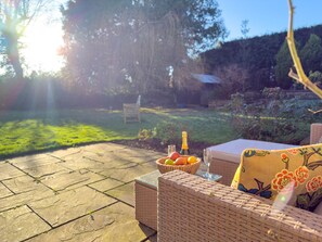 An outdoor seating area with wicker furniture, a table with a bowl of fruit and a bottle, and a view of a garden and trees. - Eden Mount, Wetheral -  a cottage that sleeps 10 guests  in 5 bedrooms (Wetheral)