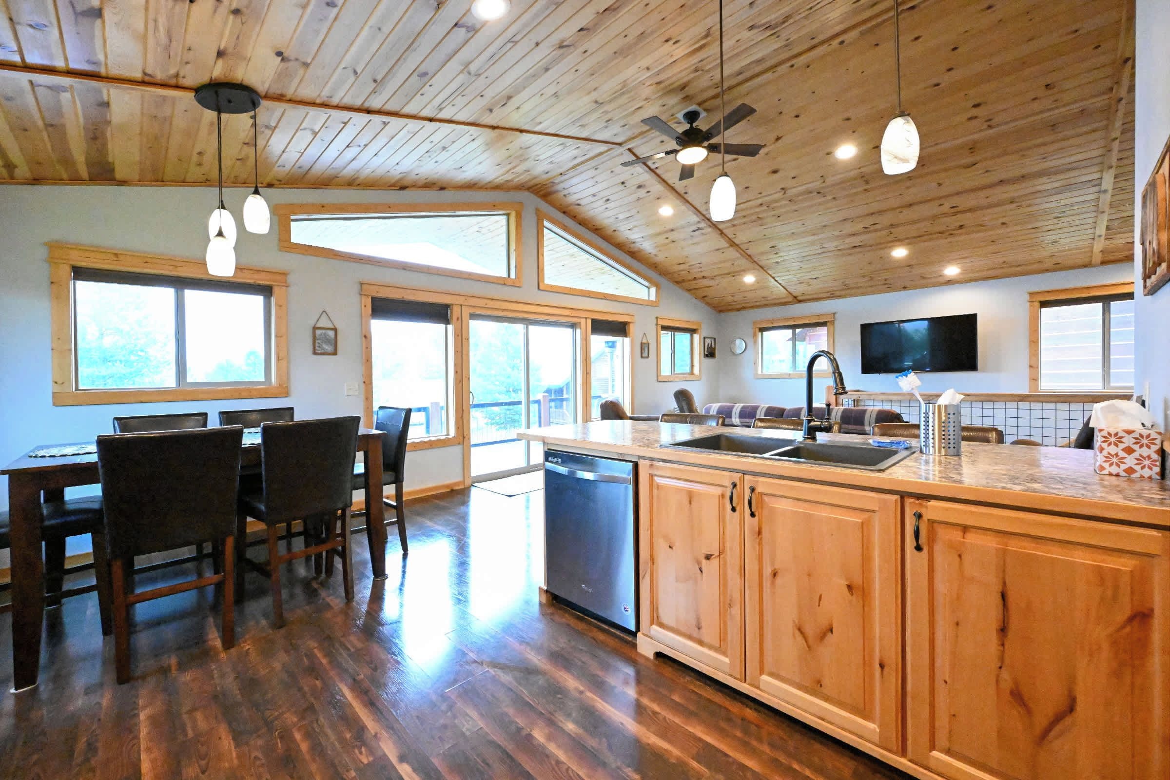 A spacious kitchen with wooden cabinets, a dishwasher, and a large island with a sink. There are multiple windows providing natural light and a ceiling fan.