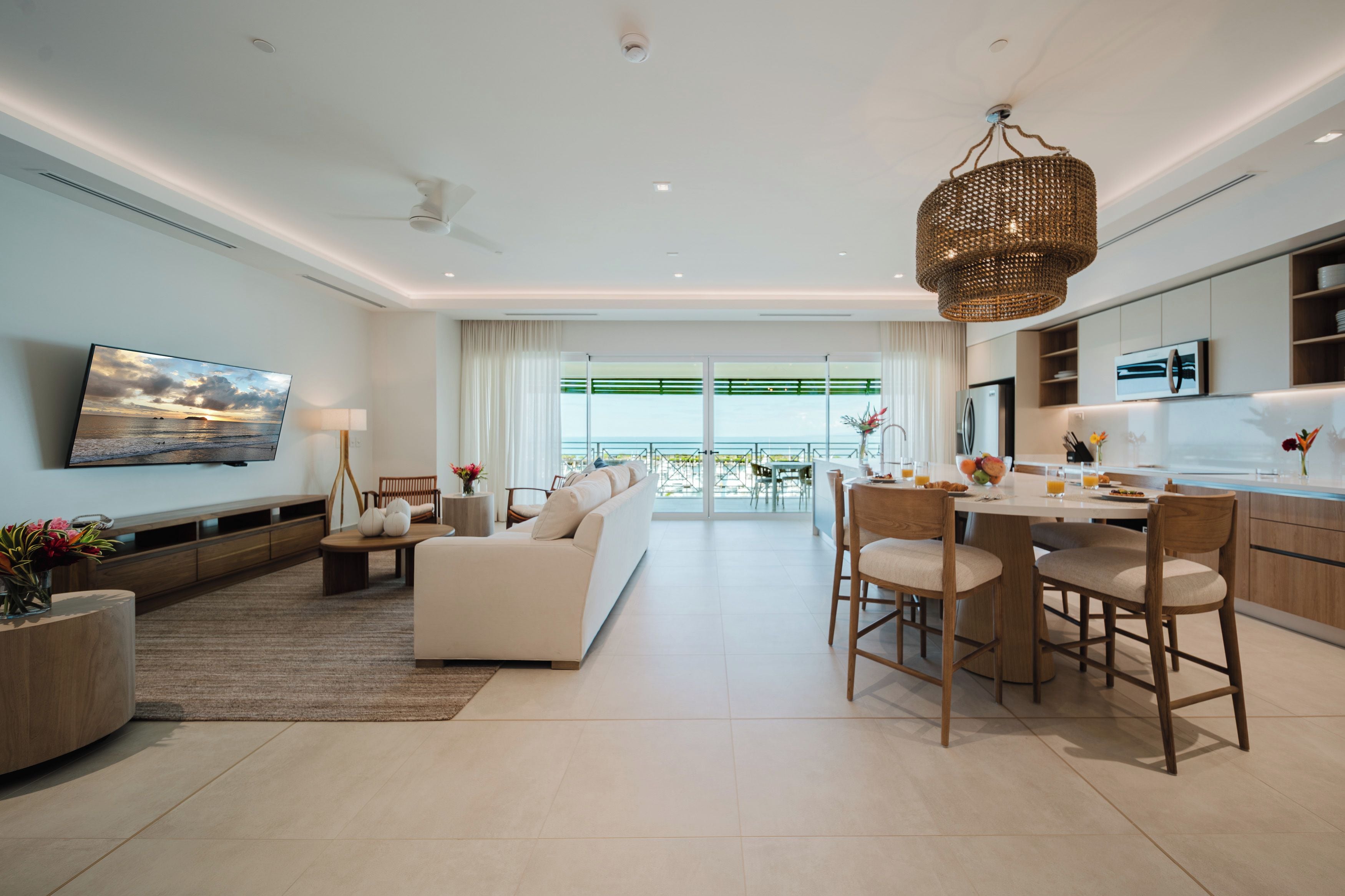 A modern living room with a sofa, dining table, and chairs, a kitchen area, and a large window with a view of the sea.