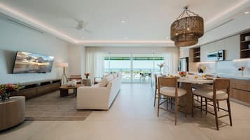 A modern living room with a sofa, dining table, and chairs, a kitchen area, and a large window with a view of the sea.