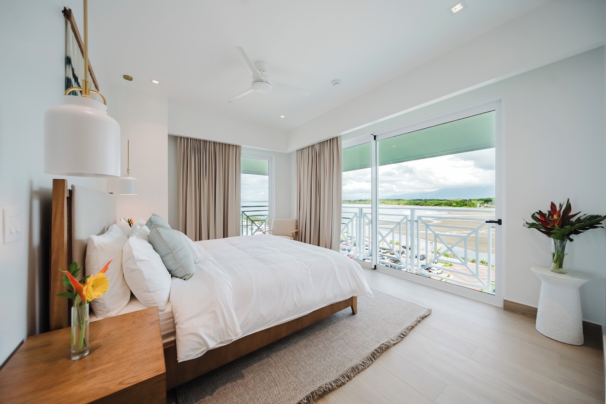 A modern bedroom with a large bed, a view of the outdoors, and a balcony with a railing.
